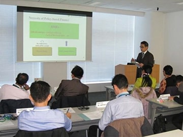 会議でスライドを説明する進行者と手元の資料を見る参加者